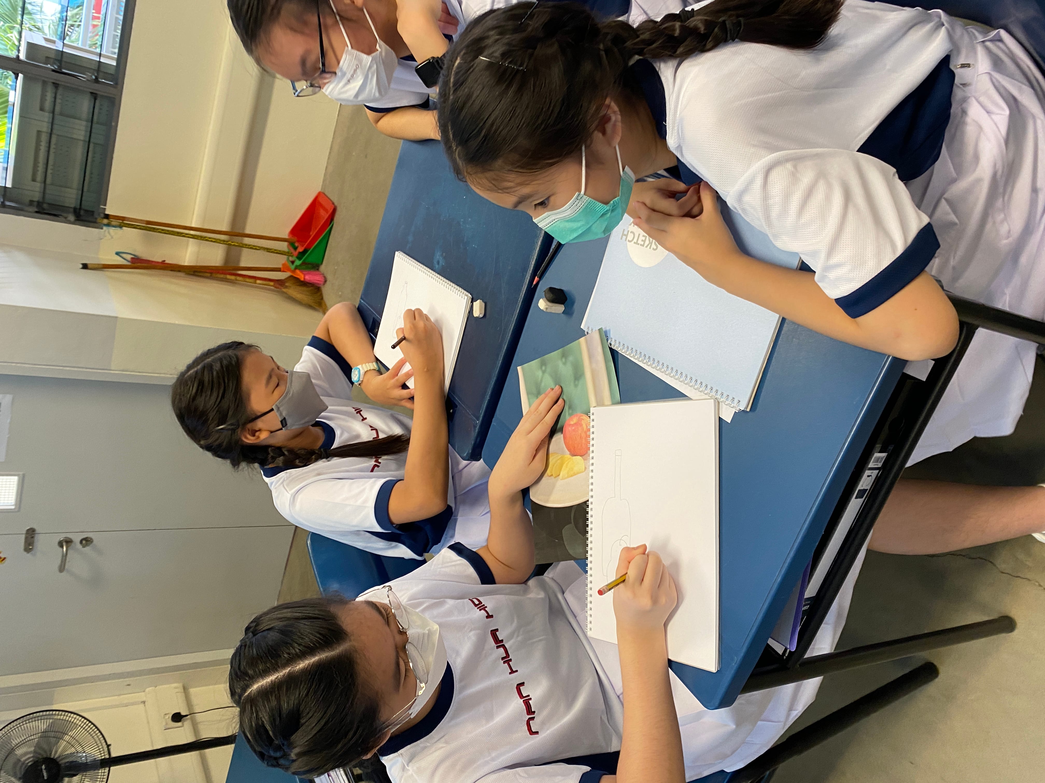 Four masked students in white uniforms sit around a blue table, sketching in notebooks during an art class.